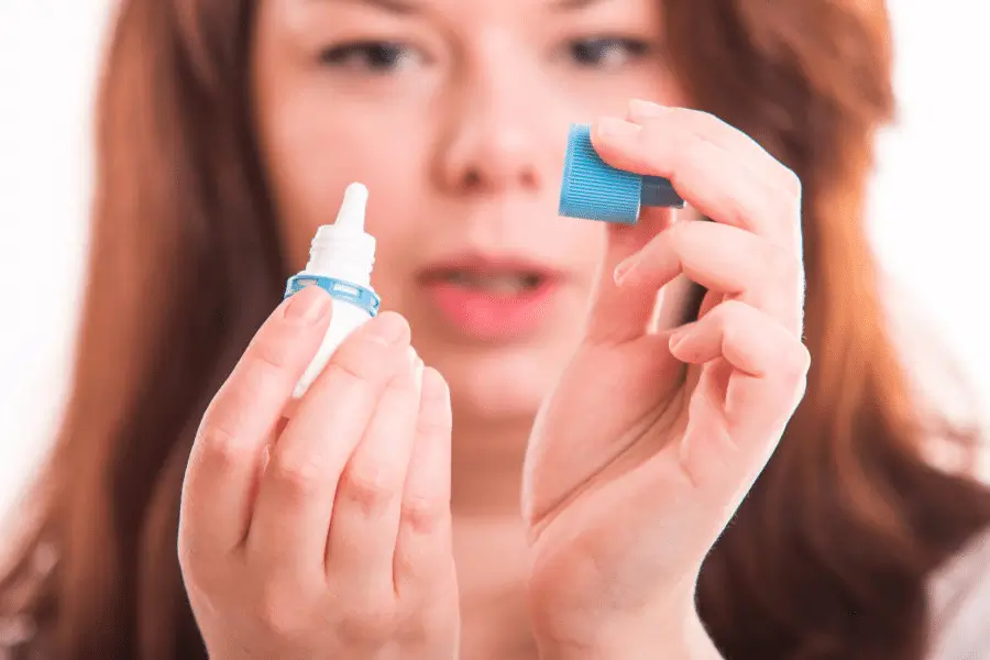 A woman holds an open eye drop bottle in one hand and the blue cap in the other, preparing to use the eye drops.
