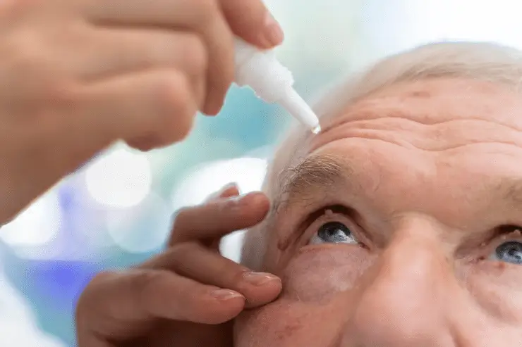 An elderly person looks up while someone applies eye drops to their right eye using a small white bottle. The image focuses on the person's face and the hand administering the drops.