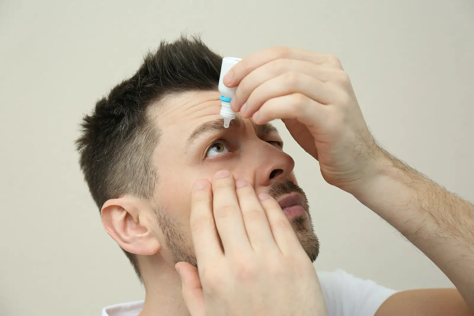 A man with short dark hair tilts his head back and uses one hand to hold open his eye while applying eye drops with the other hand.