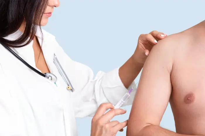 A healthcare professional wearing a white coat and stethoscope administers a Xolair injection to a man's upper arm. The man's torso is bare, and the background is light blue.