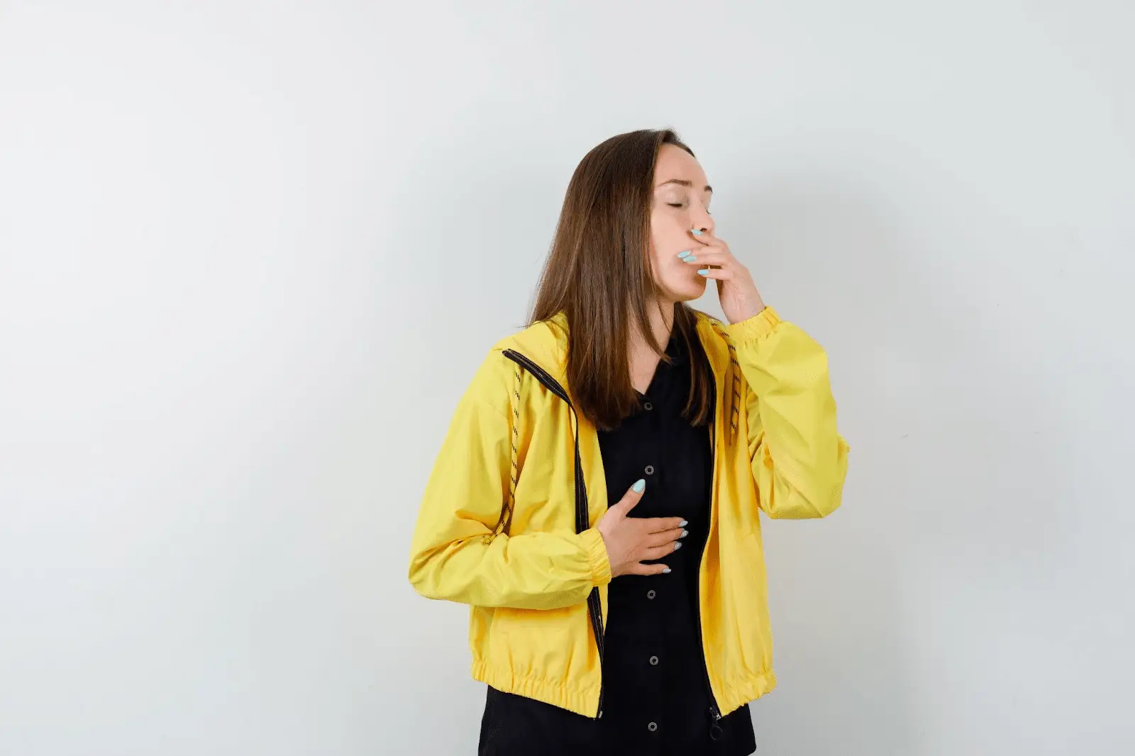A woman wearing a yellow jacket holds her stomach with one hand and covers her mouth with the other, appearing to feel nauseous or unwell, against a plain white background.