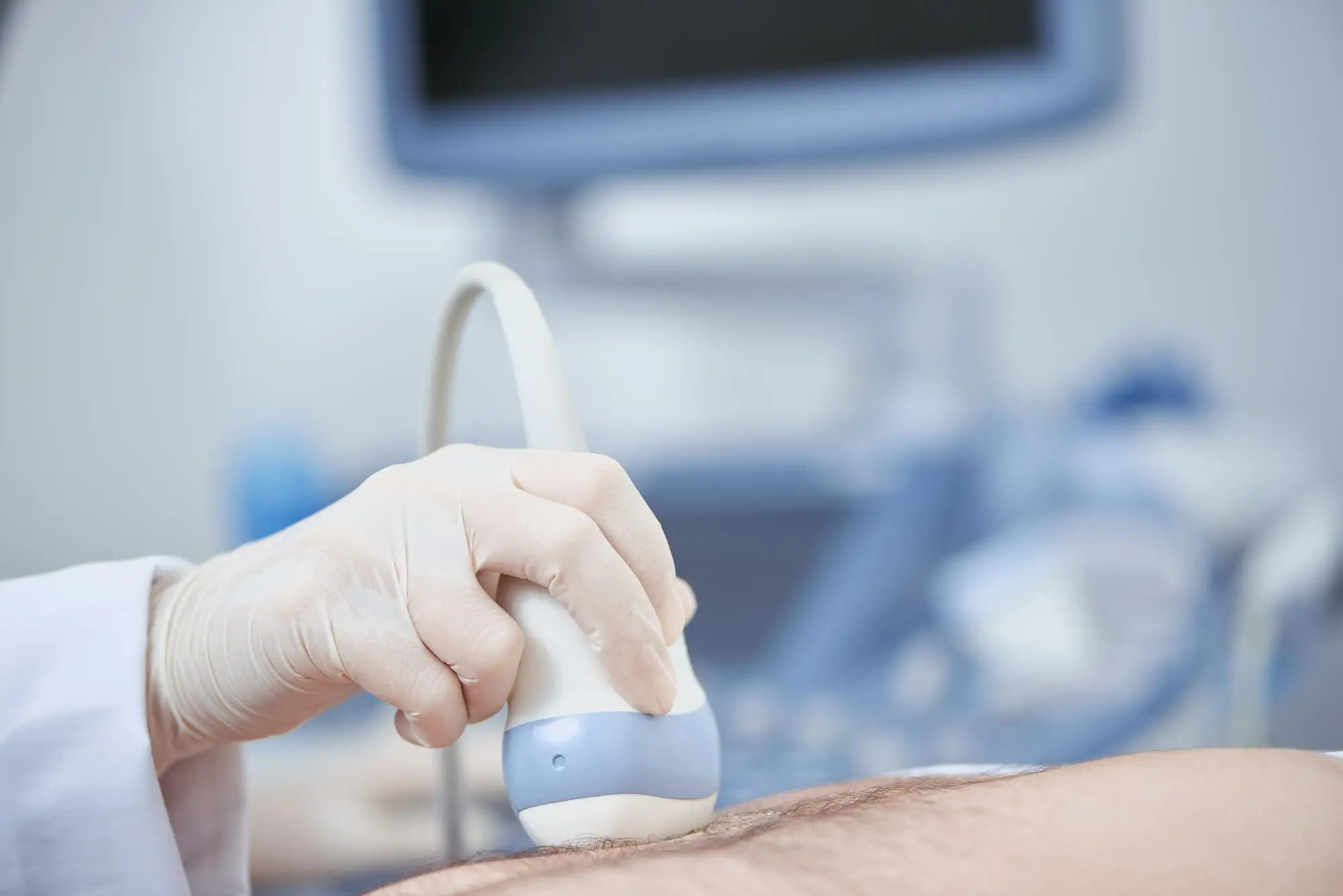 A gloved hand holds an ultrasound probe against a patient’s skin, performing a medical scan. In the background, an ultrasound machine screen is visible but out of focus.