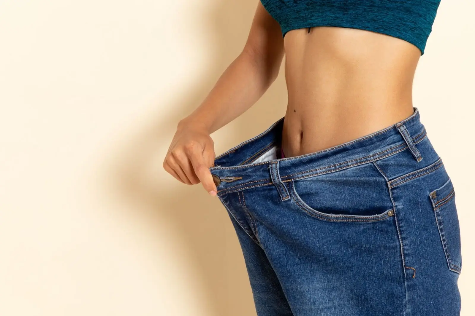 A person in a teal crop top pulls the waistband of loose blue jeans away from their waist, revealing significant weight loss. Appears against a plain beige background.