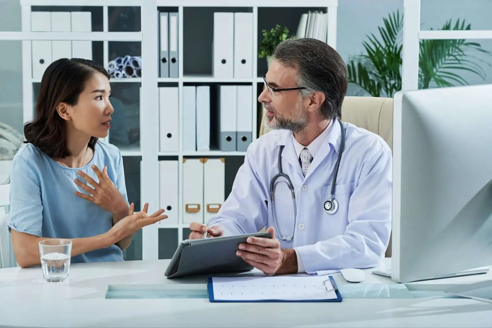 A woman sits across from a doctor in an office, gesturing with her hand while speaking. The doctor, holding a tablet and wearing a stethoscope, listens attentively. Office shelves and plants are in the background. She is asking about Mounjaro. 