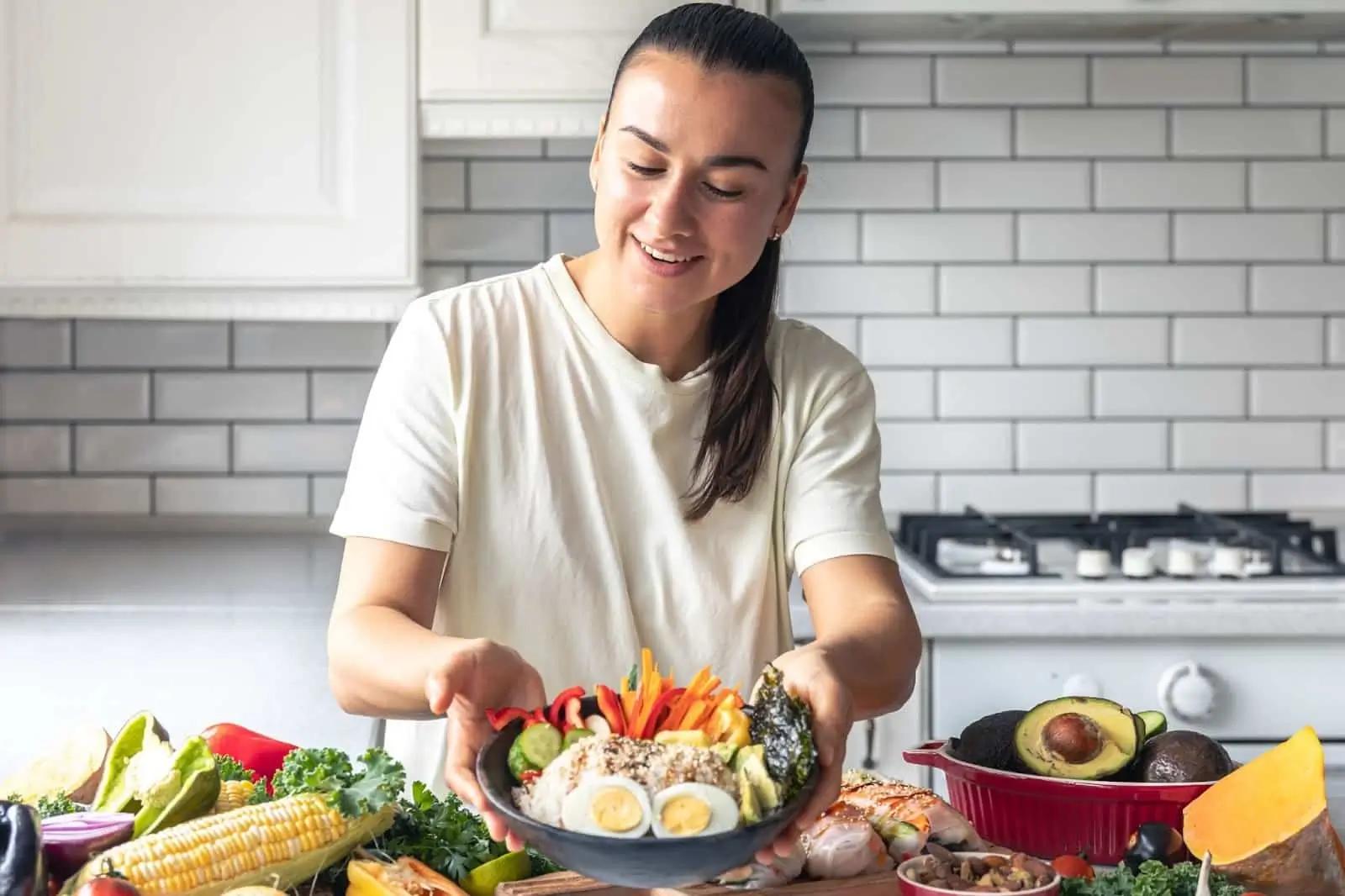 A person in a white shirt smiles while holding a bowl of colorful vegetables and grains in a kitchen. She is choosing to eat healthy during her Mounjaro treatment.