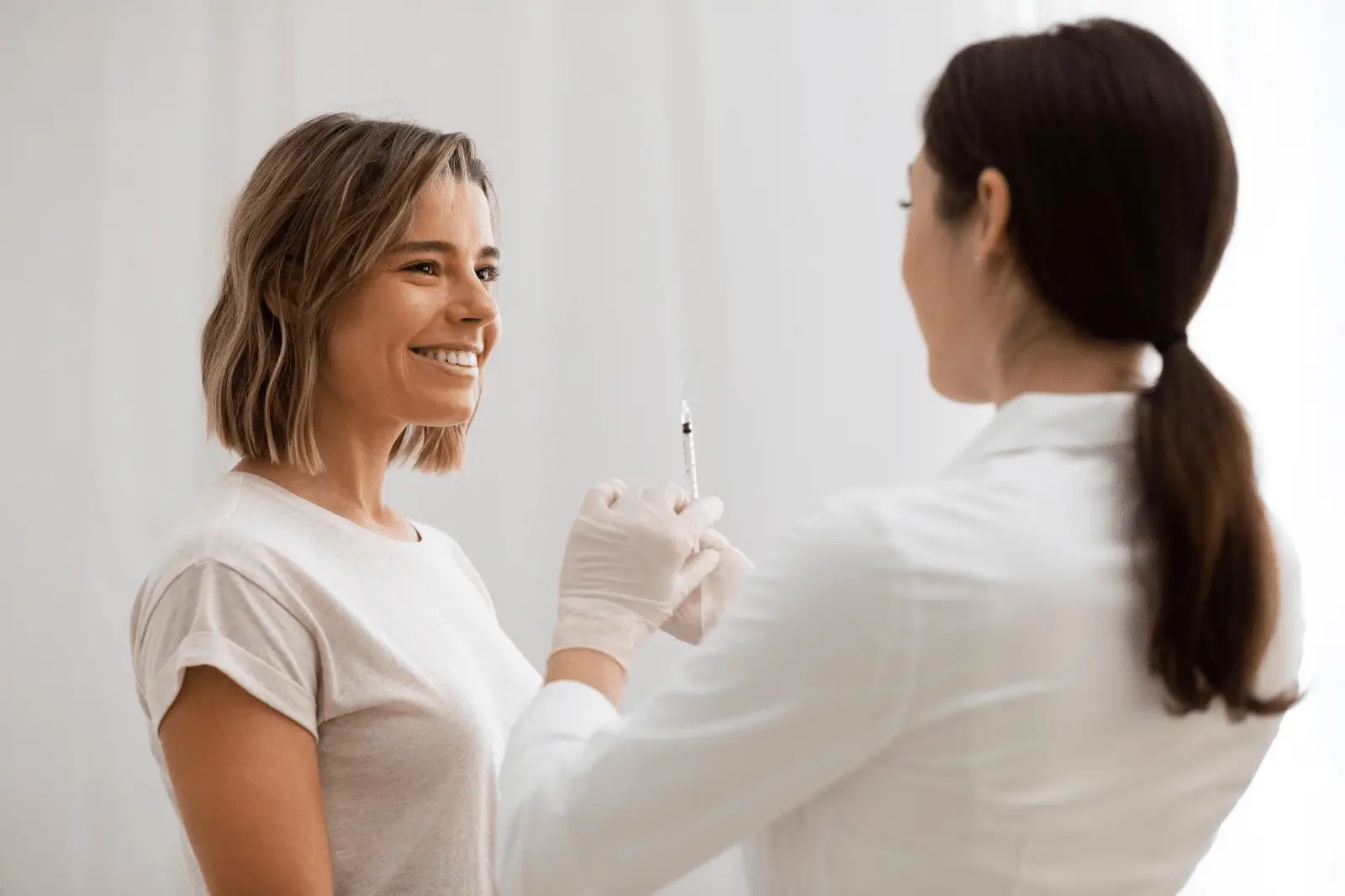 A woman in a white shirt smiles while a healthcare professional wearing gloves prepares a Xolair syringe, possibly for an injection, in a bright, clinical setting.