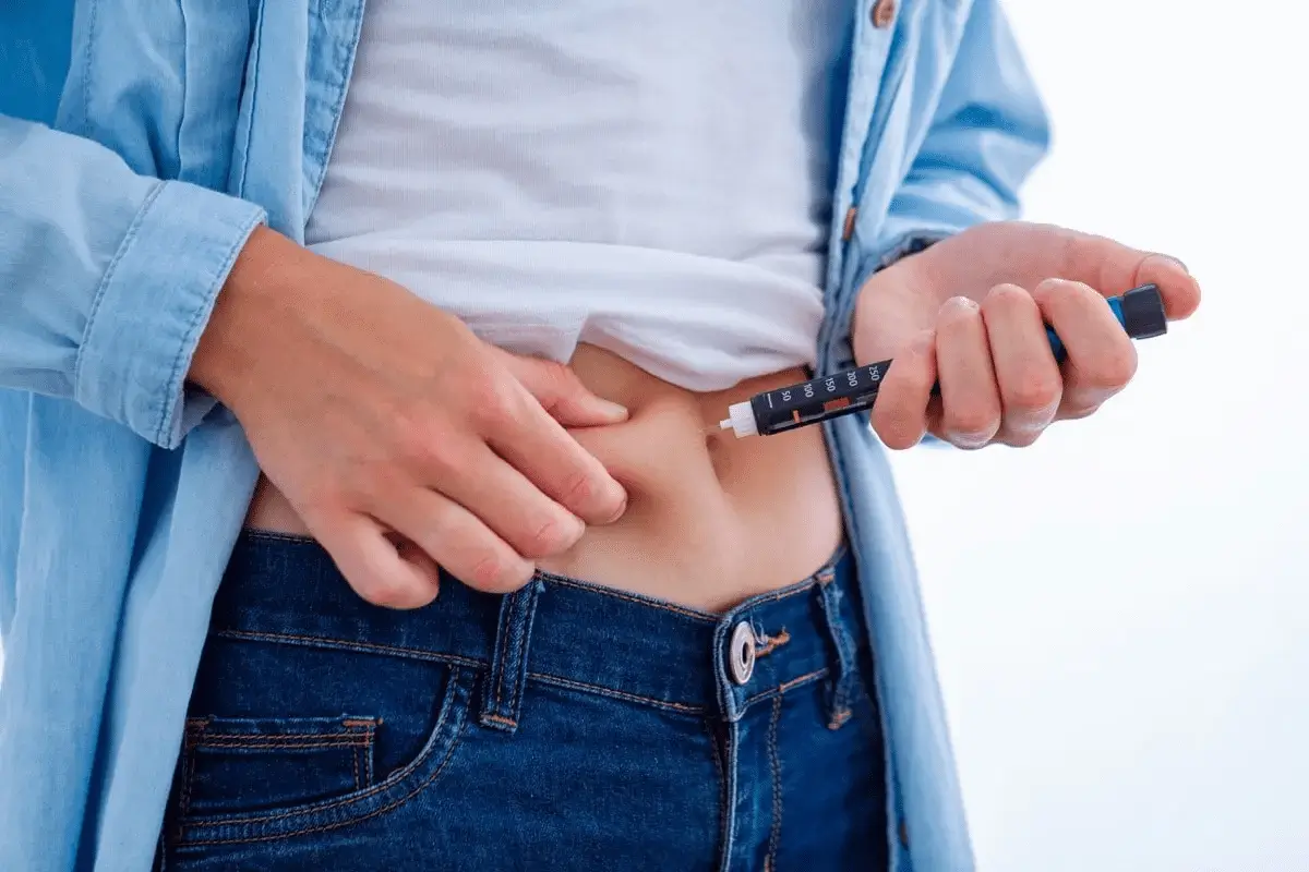 Person wearing blue jeans and a light blue shirt is injecting themselves with a Mounjaro injection in the abdomen, likely administering medication like Mounjaro or semaglutide. The focus is on the hands and the injection site.