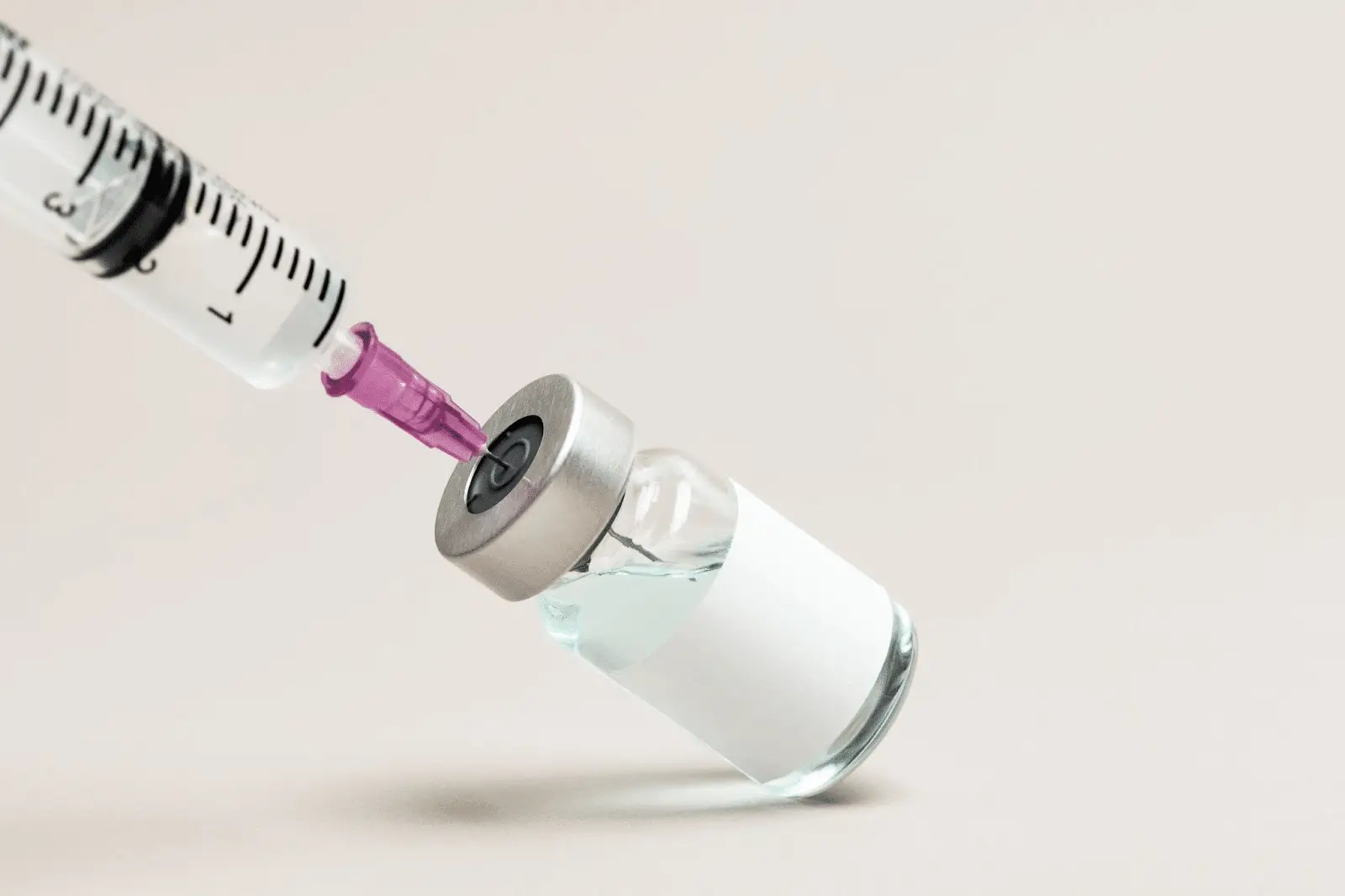 A close-up of a syringe with a purple needle drawing clear liquid from a small glass vial with a blank white label, set against a light, neutral background.