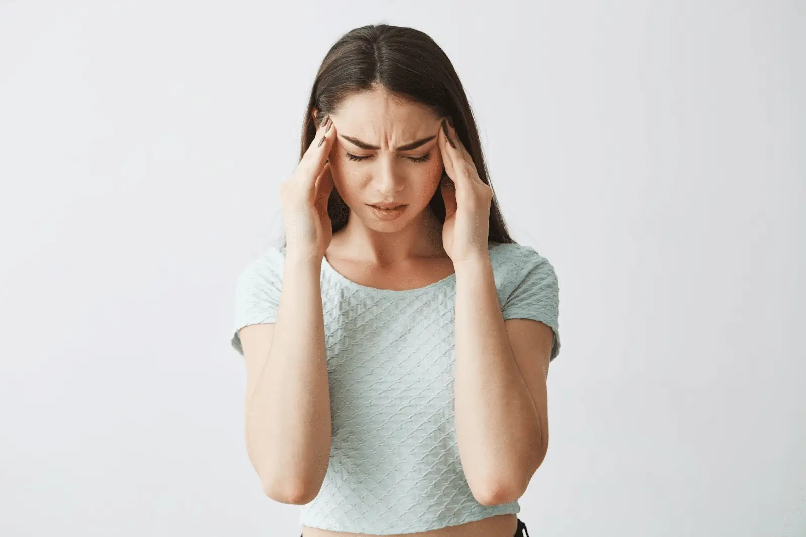 A young woman with long brown hair touches her temples with both hands and closes her eyes, appearing to be in pain or discomfort, possibly experiencing a headache due to Xolair treatment. She wears a light blue, textured top against a plain background.