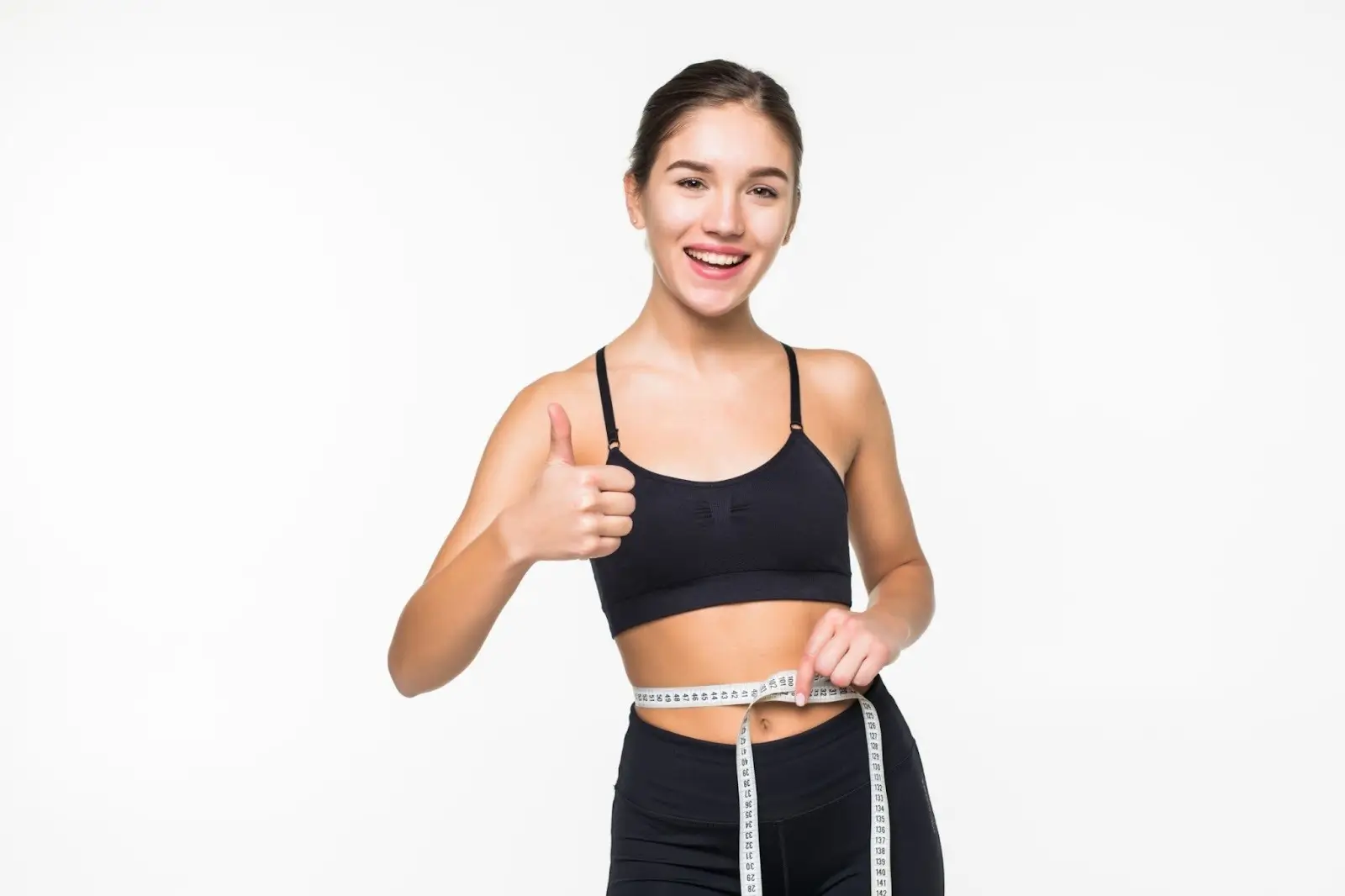 A smiling young woman in black sportswear gives a thumbs up while measuring her waist with a tape measure, standing against a plain white background, suggesting fitness or weight loss success after Mounjaro treatment.
