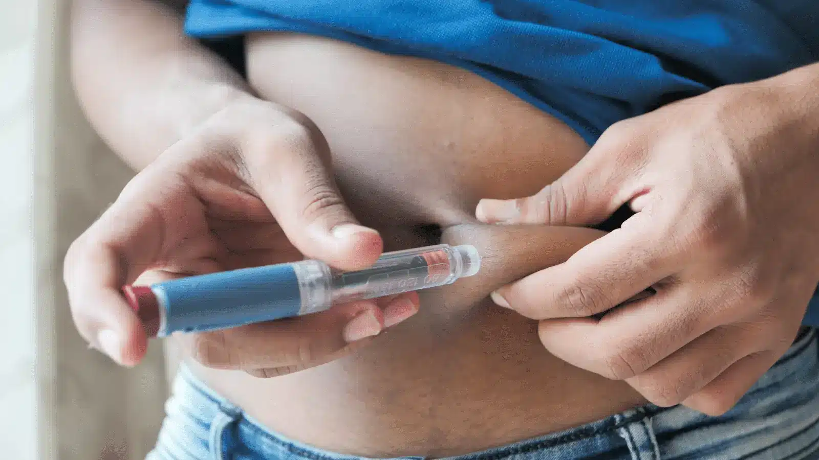 A person wearing a blue shirt injects themselves in the abdomen with a Mounjaro pen, pinching the skin with one hand and holding the injection device with the other.