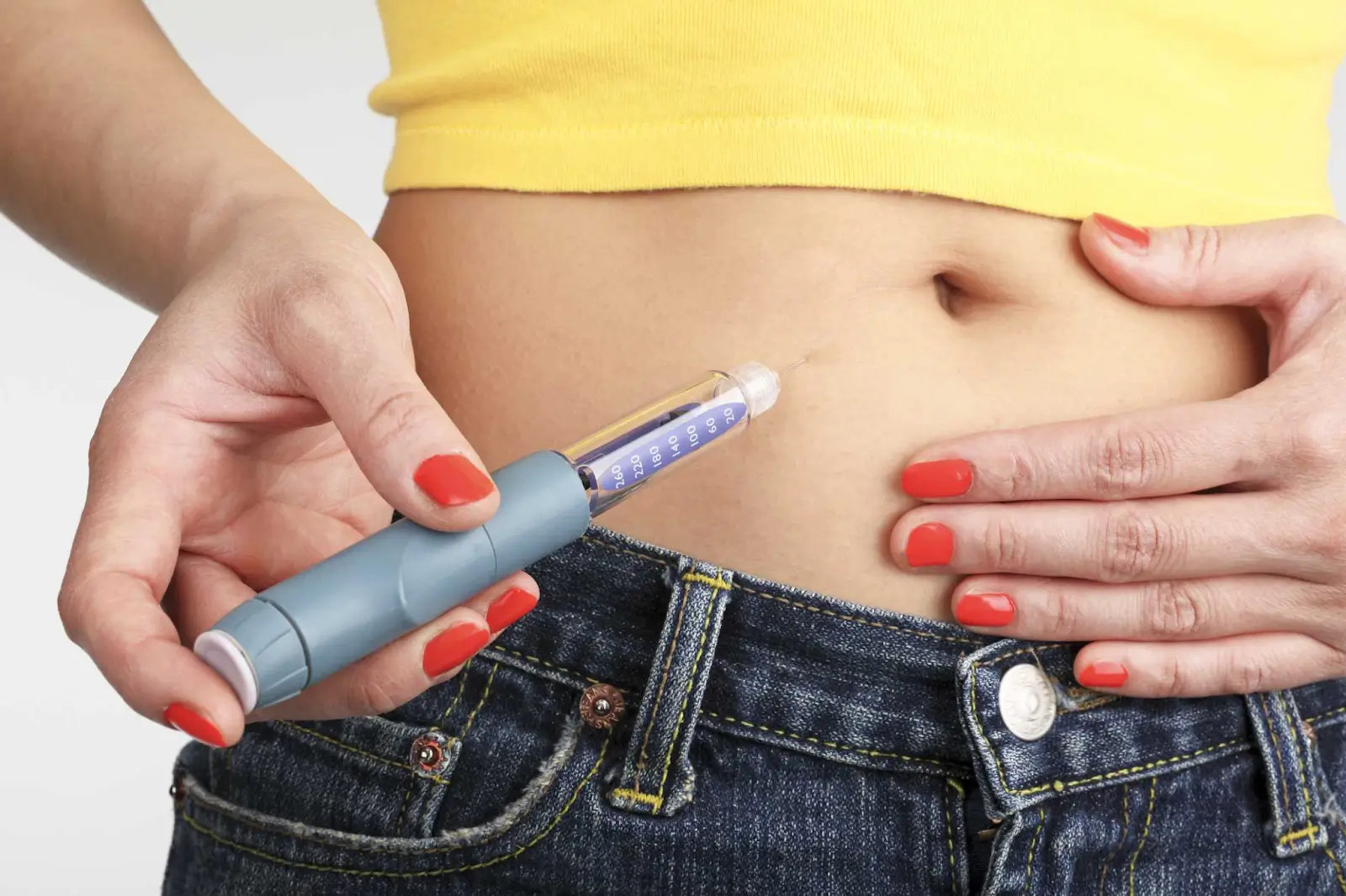 A person with red-painted nails holds a Mounjaro injection and injects it into their abdomen, with their shirt lifted and jeans slightly unbuttoned, suggesting self-administered medication.
