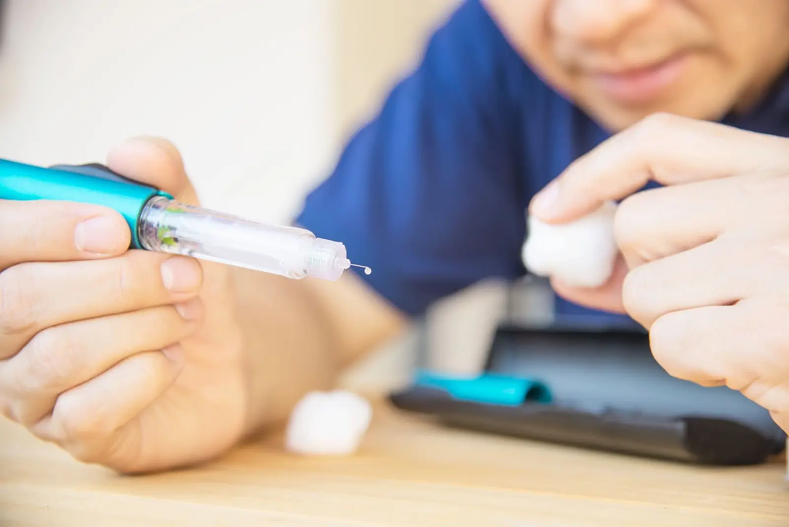 A person prepares a Mounjaro pen for injection, holding the pen with a droplet at the needle tip and a cotton ball in the other hand, with a carrying case on the table.