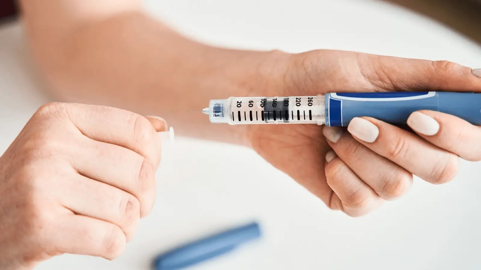 A person holding a Mounjaro pen while removing its cap, preparing for an injection. The pen is marked with black measurement lines and numbers. A blue cap lies on the white surface below.