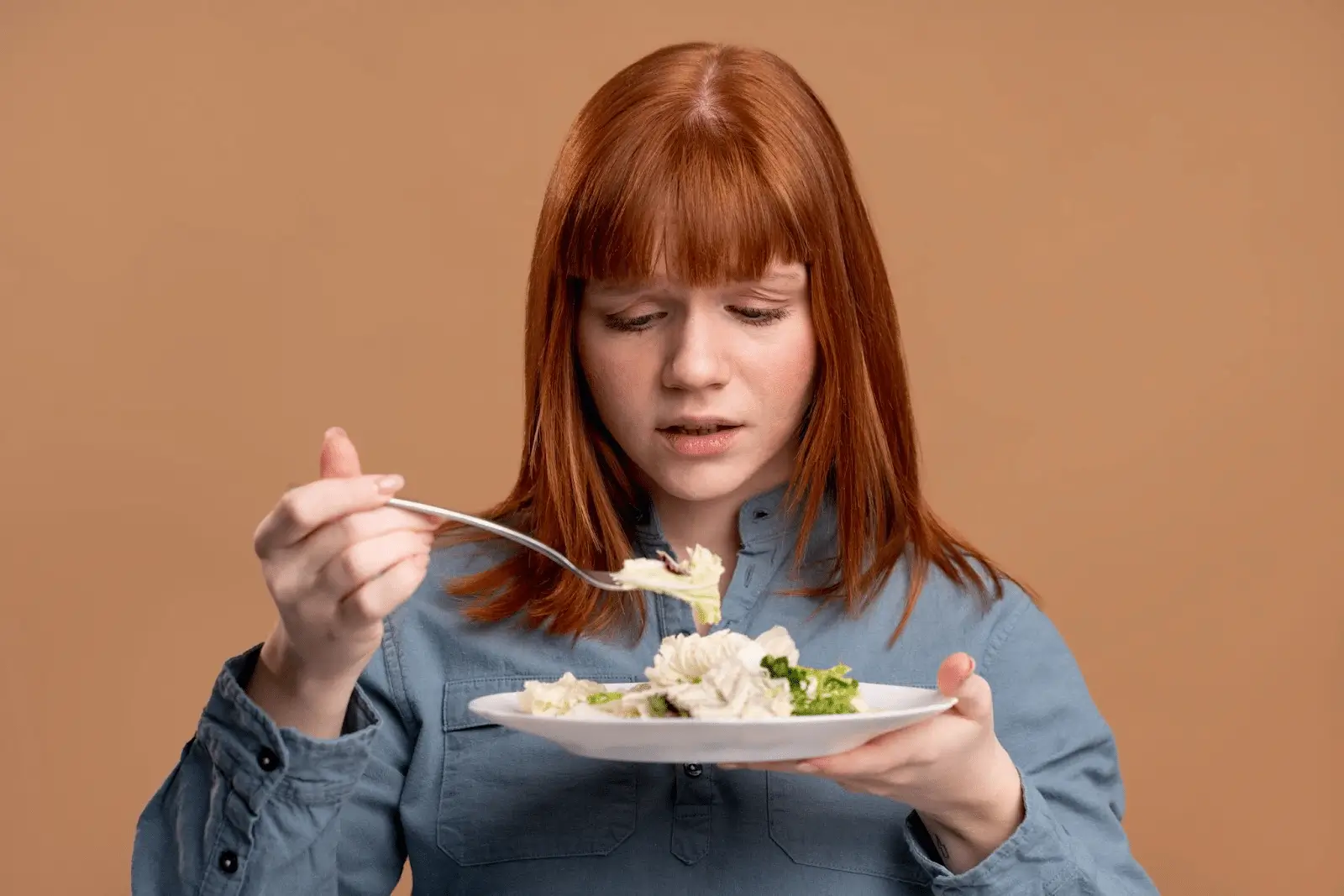 A young woman with red hair, wearing a blue button-up shirt, looks uncertainly at a forkful of pasta she is holding over a plate, set against a plain brown background.