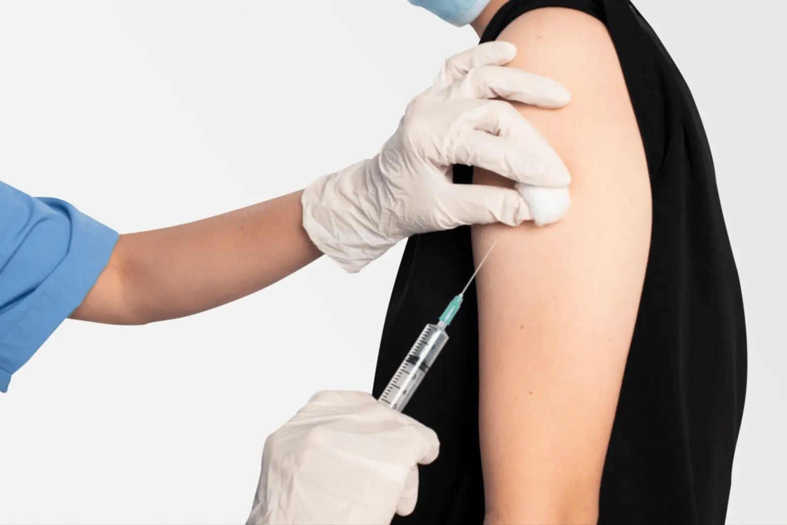 A healthcare worker wearing gloves administers a vaccine injection into the upper arm of a person wearing a sleeveless black top. The person receiving the shot is also wearing a mask.