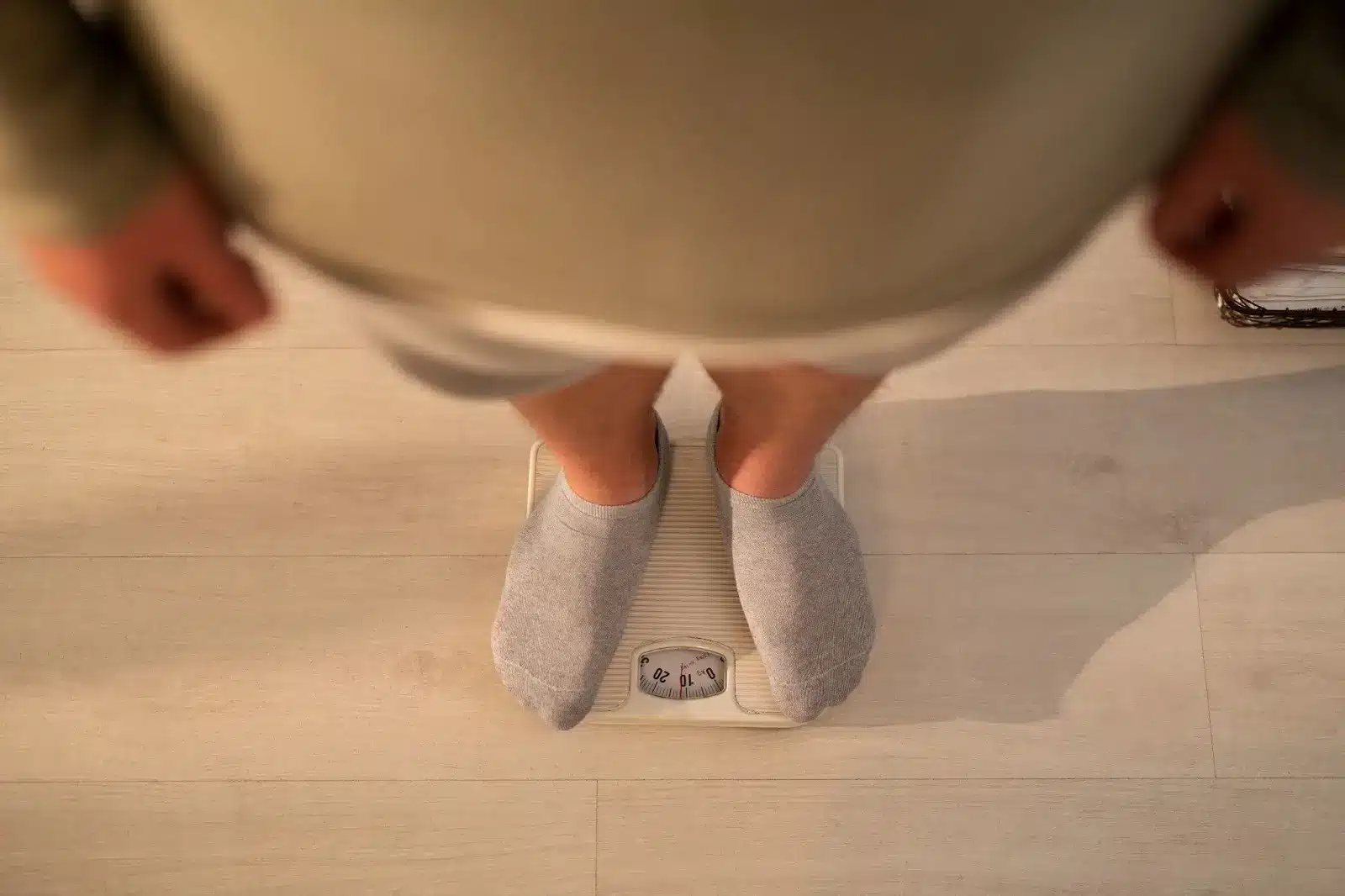 A person wearing gray socks stands on a white bathroom scale, viewed from above on a light-colored wooden floor. The scale's dial is partially visible.
