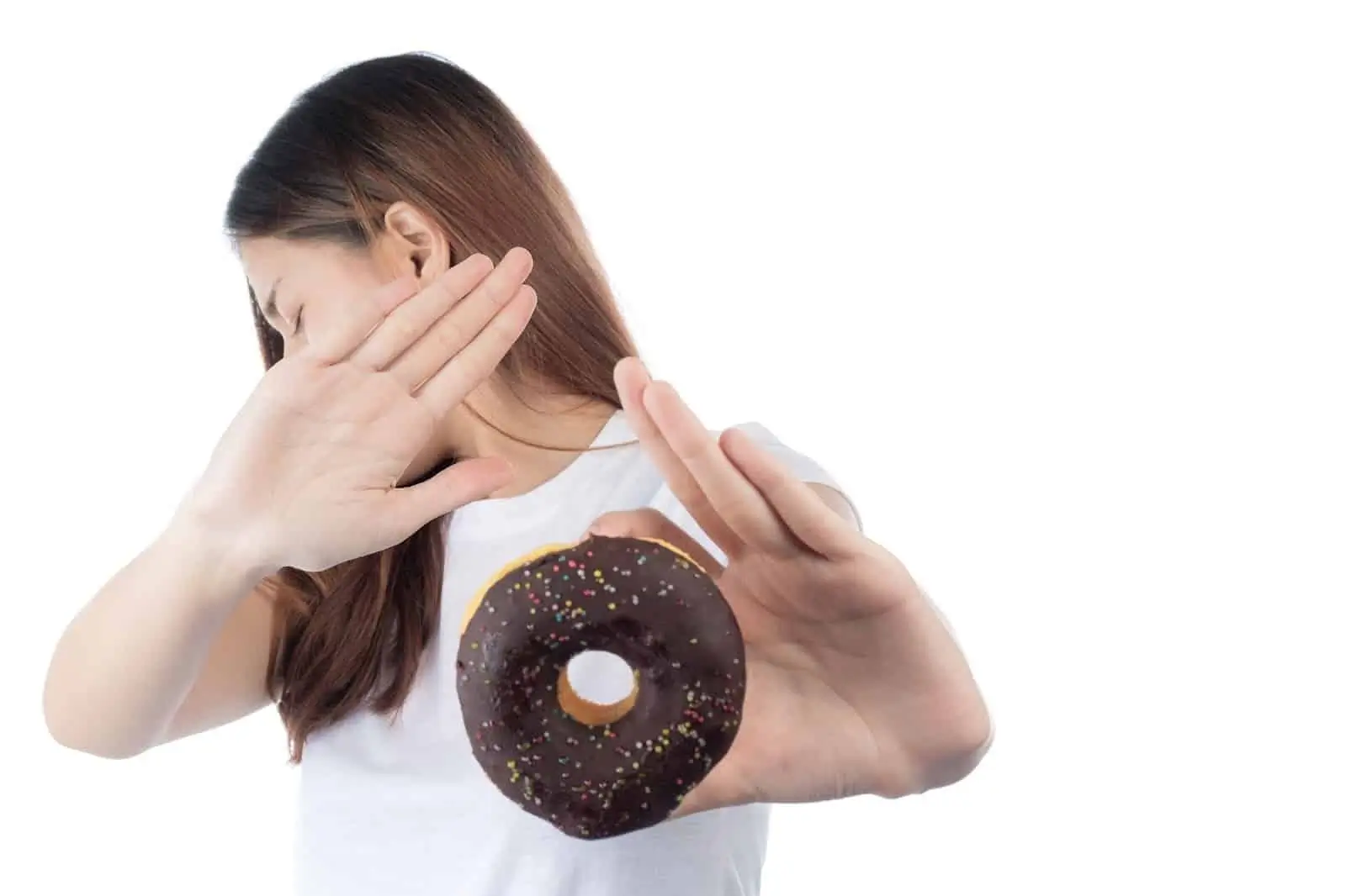 A woman in a white shirt turns away and holds up her hands to refuse a chocolate-frosted donut with sprinkles, showing a gesture of avoidance. The person in considering avoiding food after Mounjaro treatment.
