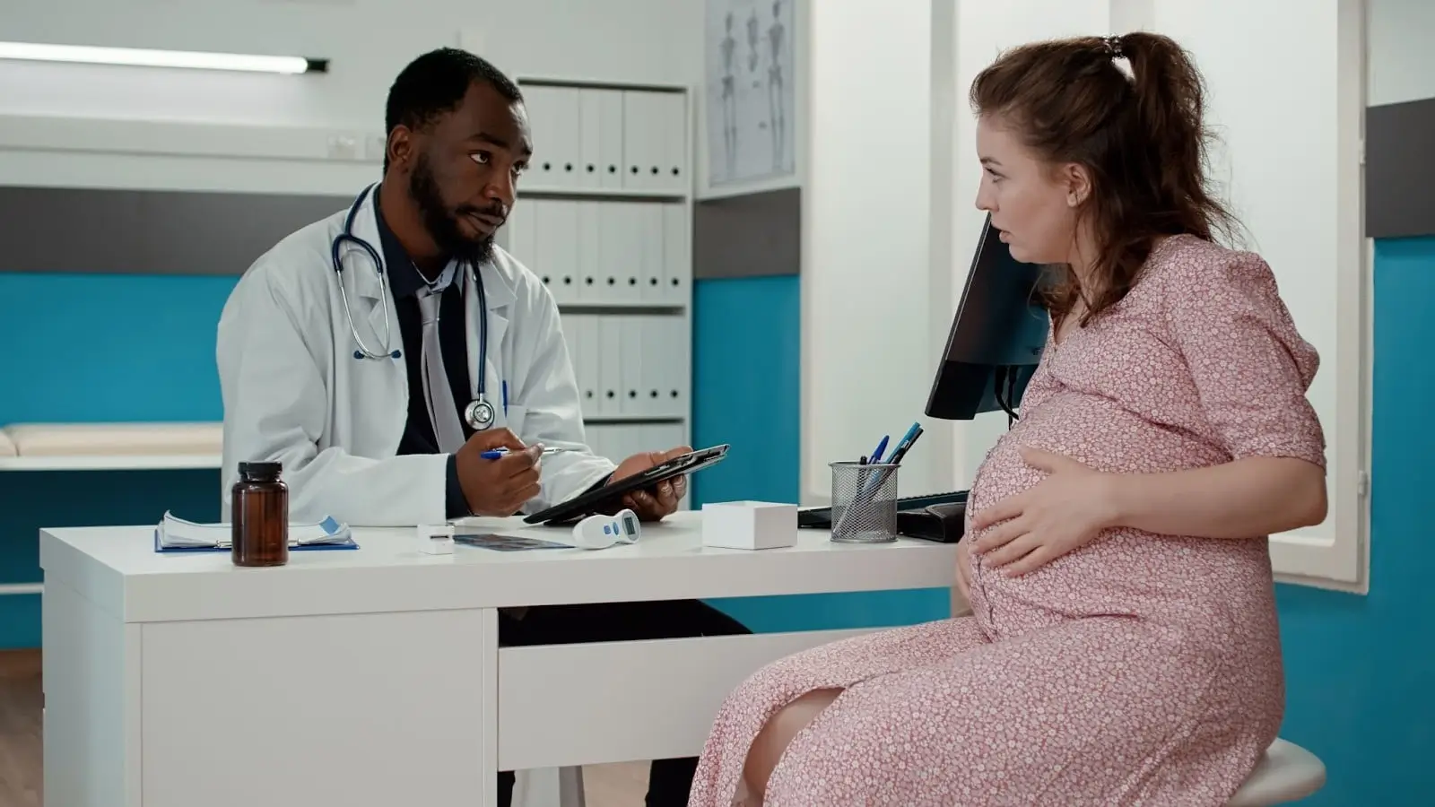 A doctor in a white coat holds a clipboard and talks to a pregnant woman in a pink dress, who is sitting and touching her belly in a medical office. She is asking about Mounjaro.