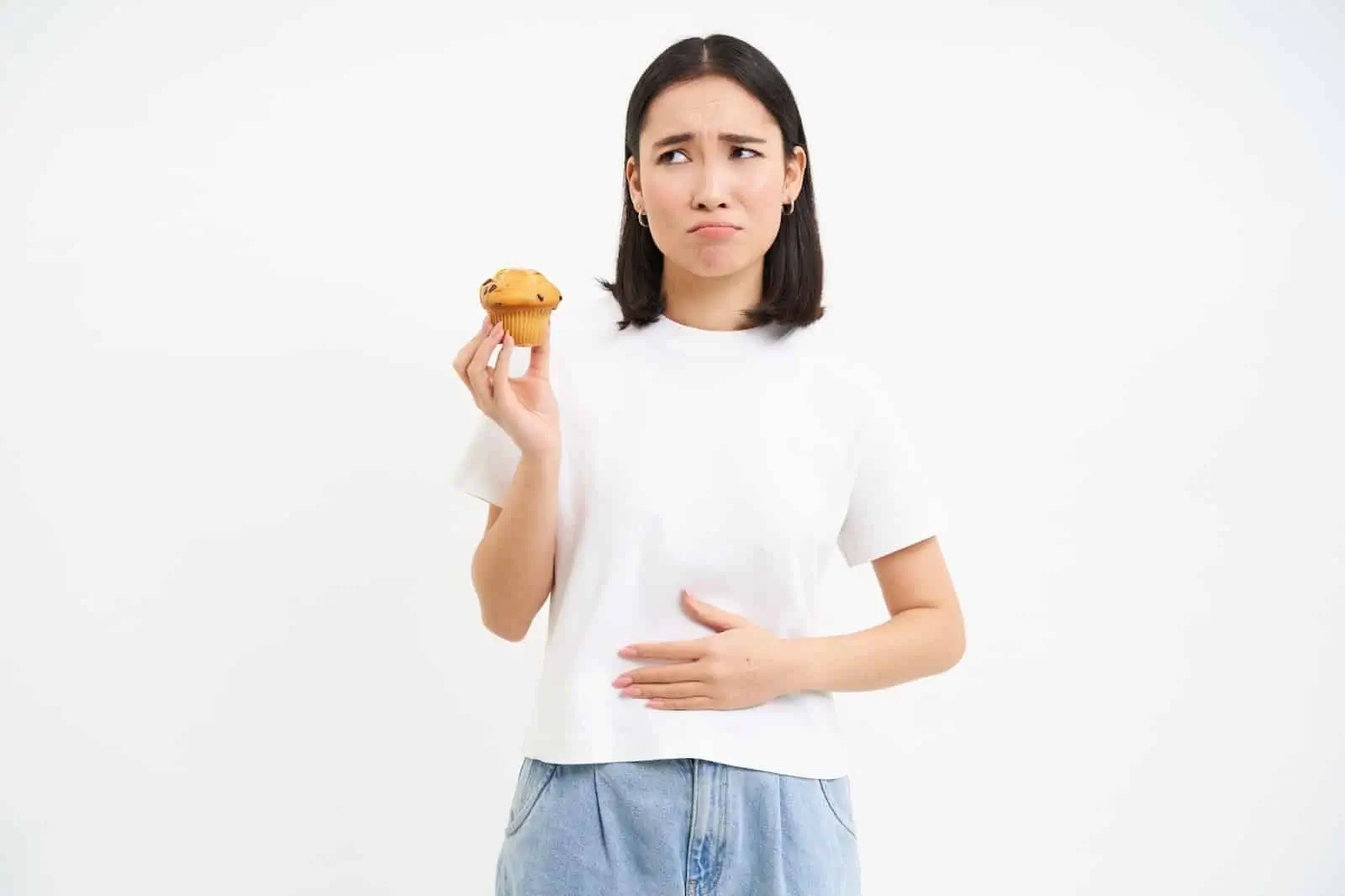 A woman in a white shirt holds a muffin in one hand and touches her stomach with the other. She feels unwell after her Mounjaro injection.