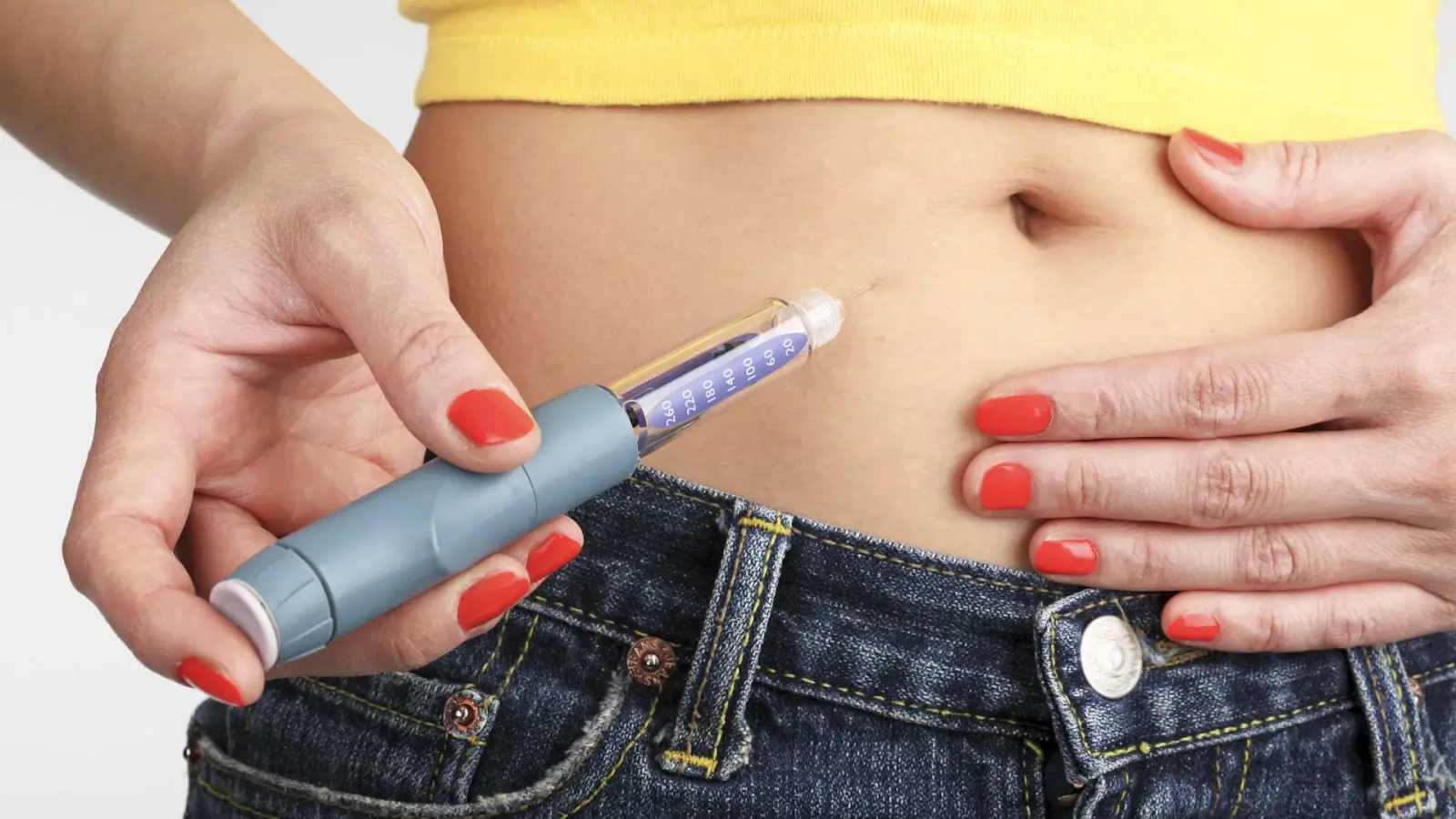A person with red-painted nails injects medication into their bare abdomen, which is one of the common Mounjaro shot locations.